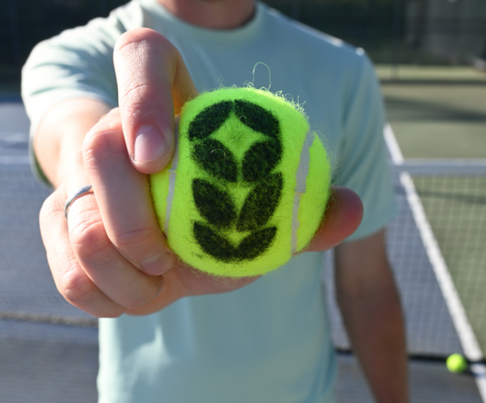 Person holding a green tennis ball with the patron logo on a tennis court